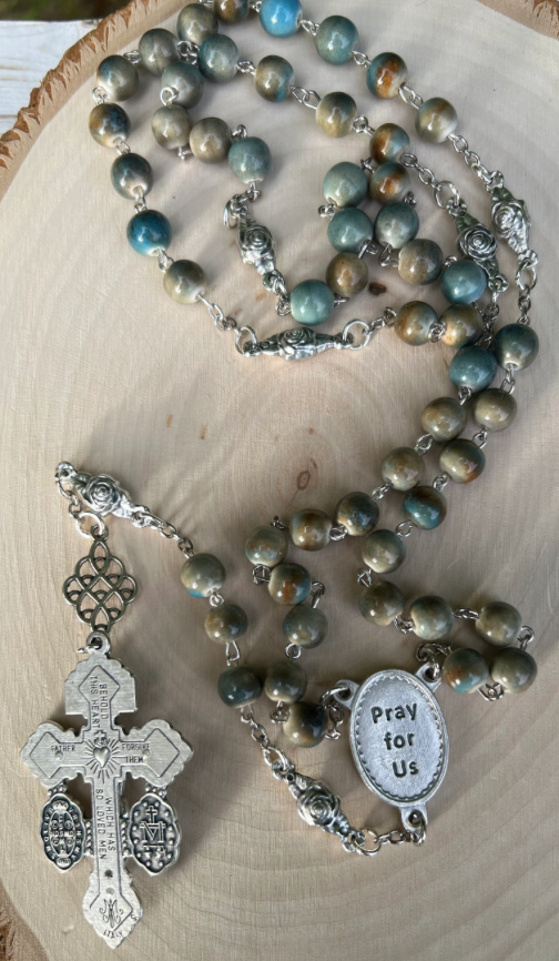 Handmade Padre Pio rosary with earth-toned ceramic beads, silver-tone chain, and a “Pray for Us” centerpiece on a light wooden background.