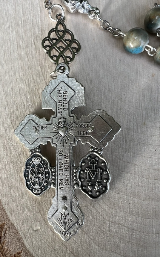 Close-up of the back of a Pardon Crucifix engraved with “Behold This Heart,” “Father Forgive Them,” and Catholic devotional medals in silver.