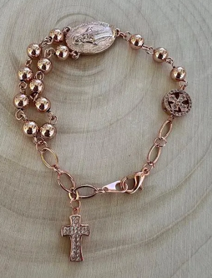 Miraculous Medal rosary bracelet with rose gold beads and cross charm displayed on natural wood background